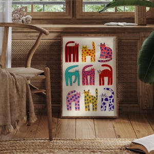 May include: A framed art print featuring nine stylized cats in various colors and patterns. The cats have red, yellow, blue, pink, and green bodies with stripes, polka dots, and floral designs. The artwork is displayed on a wooden floor.