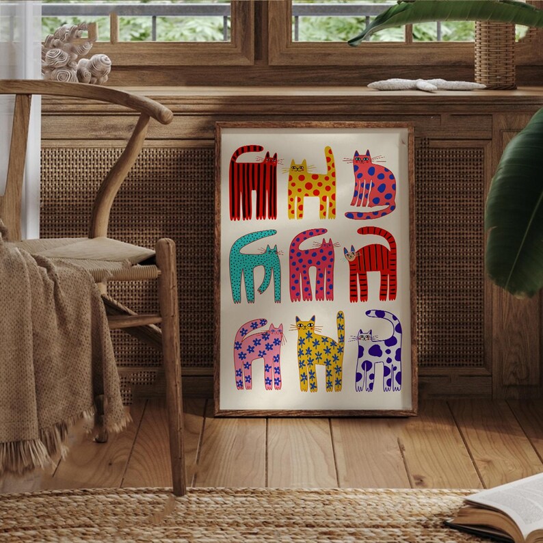 May include: A framed art print featuring nine stylized cats in various colors and patterns. The cats have red, yellow, blue, pink, and green bodies with stripes, polka dots, and floral designs. The artwork is displayed on a wooden floor.