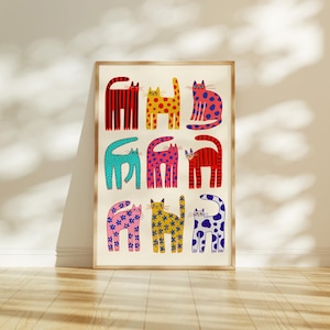 May include: A framed art print featuring nine stylized cat illustrations. Each cat is a different color and pattern, including stripes, polka dots, and floral designs. The artwork is set against a cream-colored background.