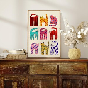 May include: A framed art print featuring nine stylized cats in various colors and patterns. The cats are arranged in three rows of three, with designs including stripes, polka dots, and floral motifs. The artwork is displayed on a wooden cabinet.