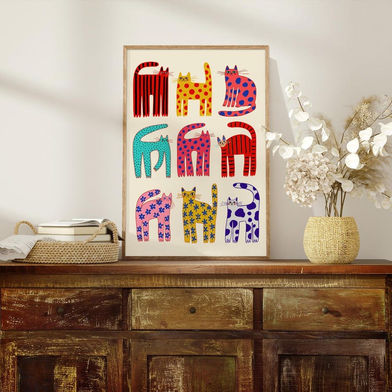 May include: A framed art print featuring nine stylized cats in various colors and patterns. The cats are arranged in three rows of three, with designs including stripes, polka dots, and floral motifs. The artwork is displayed on a wooden cabinet.
