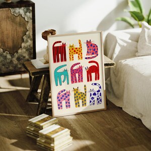 May include: A framed art print featuring a whimsical design of nine colorful cats. Each cat is uniquely patterned with stripes, polka dots, and floral designs in shades of red, yellow, blue, and pink. The artwork is displayed on a wooden surface.