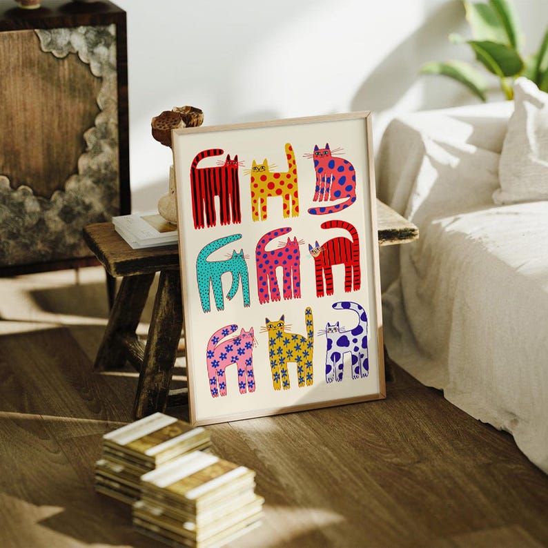 May include: A framed art print featuring a whimsical design of nine colorful cats. Each cat is uniquely patterned with stripes, polka dots, and floral designs in shades of red, yellow, blue, and pink. The artwork is displayed on a wooden surface.