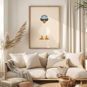 Pu&ograve; includere: Una stampa incorniciata con un'oca dei cartoni animati con berretto blu e occhiali da sole. L'oca &egrave; bianca con zampe e becco arancioni, su sfondo beige. L'opera d'arte &egrave; esposta sopra un divano color crema.