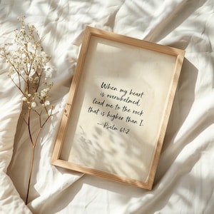 May include: A wooden framed print with the text "When my heart is overwhelmed, lead me to the rock that is higher than I. - Psalm 61:2". The print is on a cream-colored background. A sprig of white flowers is to the left.