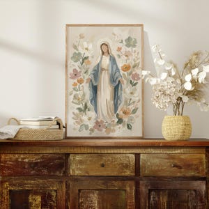 May include: A framed print of a religious figure surrounded by flowers. The figure is depicted in a long white dress and blue shawl, with a halo. The floral arrangement includes pastel colors like pink, orange, and green. The artwork is displayed on a wooden cabinet.