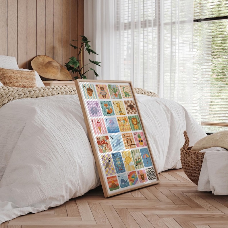 May include: A framed alphabet art print featuring colorful illustrations of animals and objects. The artwork is displayed in a light-colored wooden frame and leans against a bed with white bedding. The print includes letters A through Z.