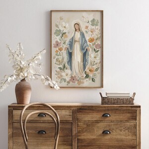 May include: A framed print of a religious figure surrounded by flowers. The figure is depicted in a long white dress and blue shawl, with a halo. The artwork has a soft, pastel color palette and is displayed on a wooden dresser.