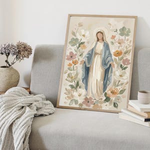 May include: A framed art print featuring a religious figure surrounded by flowers. The figure is depicted in a white dress and blue shawl, with a halo. The floral arrangement includes white, pink, and orange blooms. The artwork is set against a neutral background.