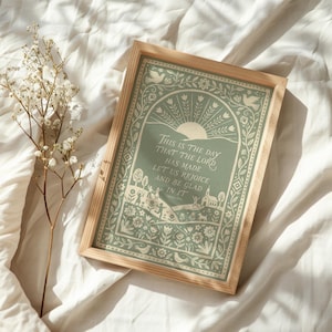 May include: A framed art print with a light green background and a wooden frame. The print features the text "THIS IS THE DAY THAT THE LORD HAS MADE LET US REJOICE AND BE GLAD IN IT" in a decorative font. The artwork includes illustrations of a sun, birds, and a landscape.