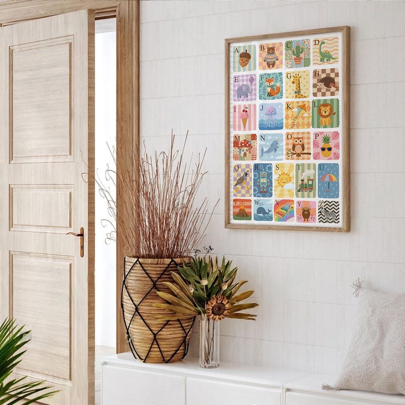 May include: A framed alphabet art print featuring colorful illustrations of animals and objects, each representing a letter. The artwork is in a wooden frame and hangs on a white wall. A decorative basket and vase with dried plants are in the foreground.