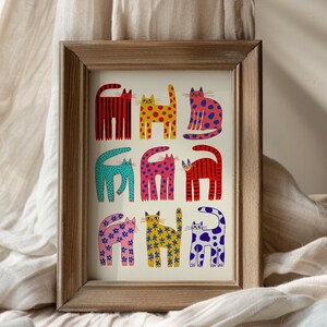 May include: A framed art print featuring nine stylized cat illustrations in various colors and patterns. The cats are arranged in three rows of three, with designs including stripes, polka dots, and floral motifs. The frame is made of light brown wood.