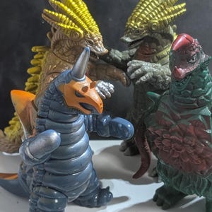 May include: Four kaiju monster action figures standing together. One is blue with an orange head crest, one is dark green with a red chest pattern, and two are textured gold and brown. A white measuring tape shows a scale of 20 centimeters at the base.
