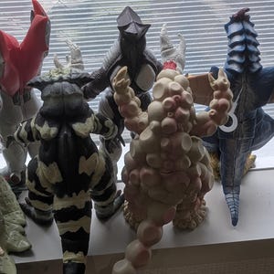 Op de afbeelding: Een verzameling van acht staande monsteractiefiguren, gezien vanaf de achterkant. De figuren hebben diverse texturen, waaronder gladde, hobbelige en geschubde oppervlakken, met kleuren vari&euml;rend van beige, zwart en wit tot rood en blauw.