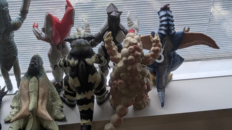 Op de afbeelding: Een verzameling van acht staande monsteractiefiguren, gezien vanaf de achterkant. De figuren hebben diverse texturen, waaronder gladde, hobbelige en geschubde oppervlakken, met kleuren vari&euml;rend van beige, zwart en wit tot rood en blauw.