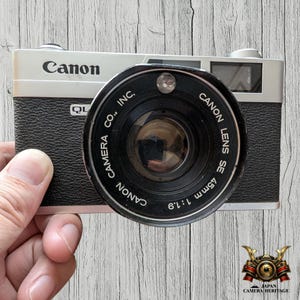 May include: A vintage Canon QL19 camera with a black body and silver top. The lens is black with white text, including "Canon Lens SE 45mm 1:1.9". The camera is held by a hand against a wooden background.