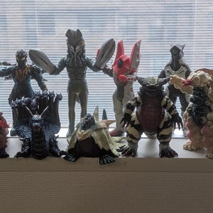 Op de afbeelding: Een verzameling van elf diverse monsteractiefiguren die in twee rijen staan opgesteld. De figuren hebben uiteenlopende ontwerpen, waaronder wezens met vleugels, meerdere koppen, hoorns en een textuurrijke huid in kleuren zoals bruin, zwart, rood, wit en blauw.