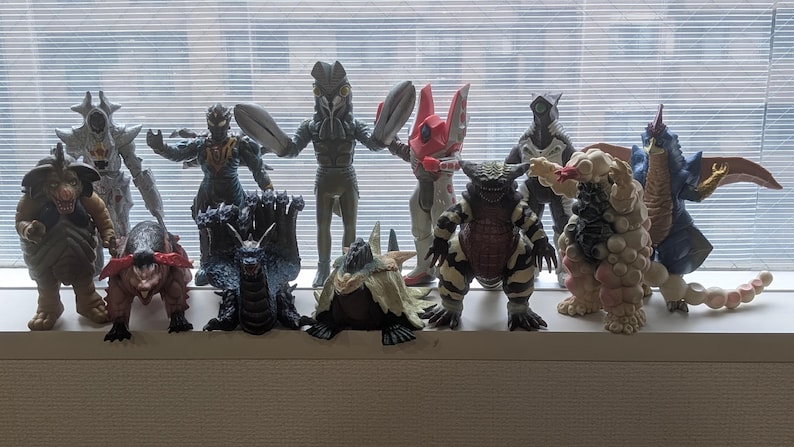 Op de afbeelding: Een verzameling van elf diverse monsteractiefiguren die in twee rijen staan opgesteld. De figuren hebben uiteenlopende ontwerpen, waaronder wezens met vleugels, meerdere koppen, hoorns en een textuurrijke huid in kleuren zoals bruin, zwart, rood, wit en blauw.