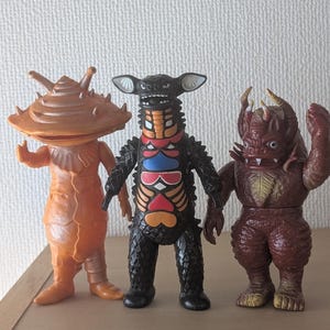 May include: Three standing monster figurines. The left figure is orange with a wide, flat, ridged head. The center figure is black with a patterned chest of red, blue, and yellow shapes. The right figure is brown with horns and a textured body.