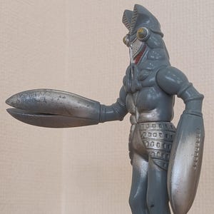 May include: A gray and silver action figure of a monster with a segmented head, claw-like hands, and a shield on one arm. The figure is standing on a wooden surface.