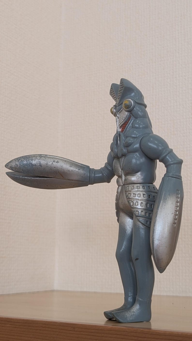 May include: A gray and silver action figure of a monster with a segmented head, claw-like hands, and a shield on one arm. The figure is standing on a wooden surface.
