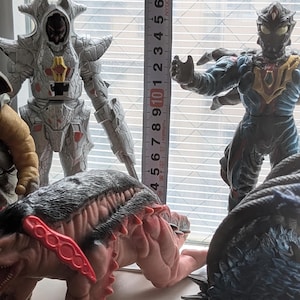 Op de afbeelding: Een verzameling van vier monsteractiefiguren geplaatst naast een verticaal meetlint. De figuren omvatten een zilveren gepantserd wezen, een donkerblauw en zwart mensachtig figuur, een beige getextureerd figuur en een roze-bruin wezen met rode stekels.