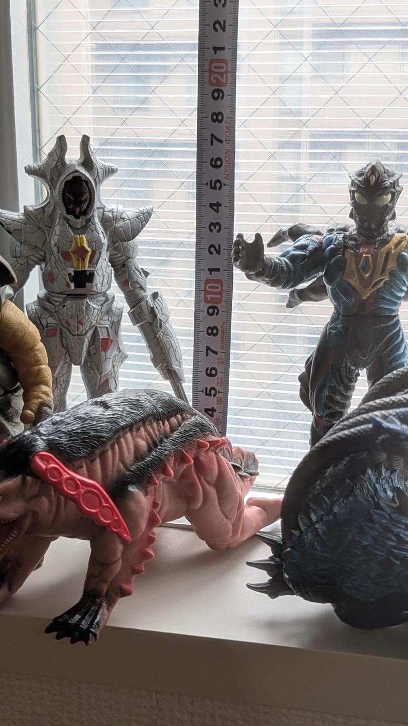 Op de afbeelding: Een verzameling van vier monsteractiefiguren geplaatst naast een verticaal meetlint. De figuren omvatten een zilveren gepantserd wezen, een donkerblauw en zwart mensachtig figuur, een beige getextureerd figuur en een roze-bruin wezen met rode stekels.