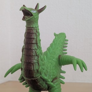 May include: A green monster figurine with a long neck, a brown textured chest, and a reptilian appearance. The creature features sharp claws, a pointed tail, and small horns on its head. Its mouth is open, revealing teeth, and it has white eyes.