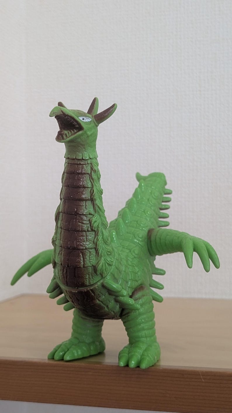 May include: A green monster figurine with a long neck, a brown textured chest, and a reptilian appearance. The creature features sharp claws, a pointed tail, and small horns on its head. Its mouth is open, revealing teeth, and it has white eyes.