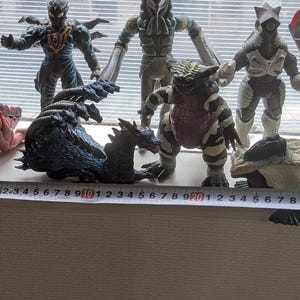 Op de afbeelding: Een verzameling van zes veelkleurige monsteractiefiguren opgesteld in een rij. Een meetlint is voor de figuren geplaatst en toont een totale breedte van ongeveer 28 centimeter, waarbij de individuele figuren in hoogte vari&euml;ren.