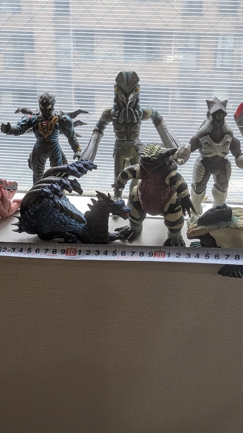 Op de afbeelding: Een verzameling van zes veelkleurige monsteractiefiguren opgesteld in een rij. Een meetlint is voor de figuren geplaatst en toont een totale breedte van ongeveer 28 centimeter, waarbij de individuele figuren in hoogte vari&euml;ren.