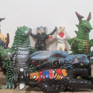 May include: A collection of various plastic monster figurines in different poses, including green, black, and grey creatures with textured skin, horns, and spikes, arranged in a group with some standing upright and others lying down.
