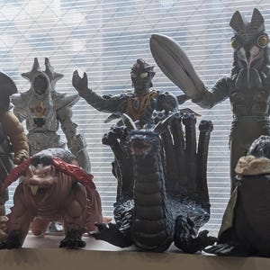 Op de afbeelding: Een verzameling van diverse plastic monsteractiefiguren die in een groep staan. De figuren hebben uiteenlopende ontwerpen, waaronder een bruin wezen, een zilveren gepantserde figuur, een donkere draak met meerdere koppen en een grote grijze figuur.