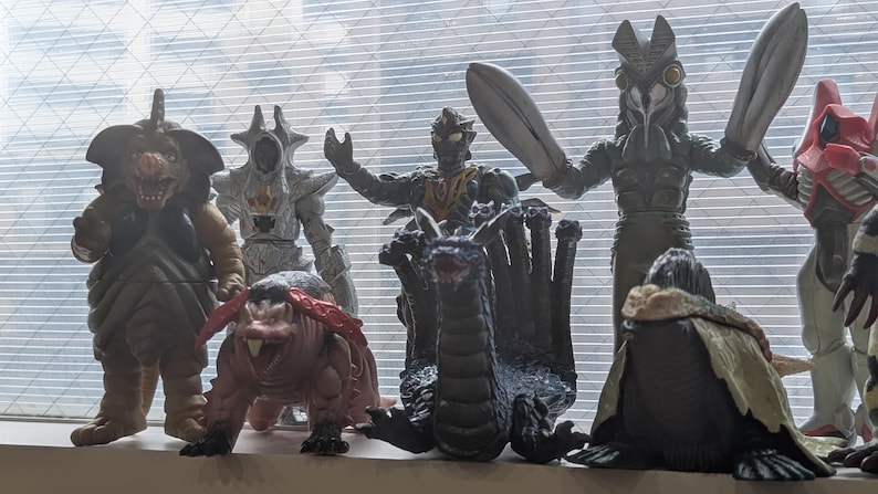 Op de afbeelding: Een verzameling van diverse plastic monsteractiefiguren die in een groep staan. De figuren hebben uiteenlopende ontwerpen, waaronder een bruin wezen, een zilveren gepantserde figuur, een donkere draak met meerdere koppen en een grote grijze figuur.