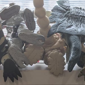 Op de afbeelding: Een verzameling van vier plastic monsteractiefiguren gezien vanaf de achterkant, met diverse texturen en kleuren waaronder zwart, wit, grijs en beige. Elk figuur toont gegoten details en fabrikantmarkeringen op de onderkant van de voeten.
