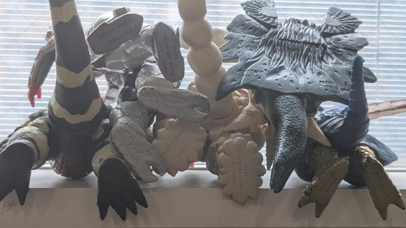 Op de afbeelding: Een verzameling van vier plastic monsteractiefiguren gezien vanaf de achterkant, met diverse texturen en kleuren waaronder zwart, wit, grijs en beige. Elk figuur toont gegoten details en fabrikantmarkeringen op de onderkant van de voeten.