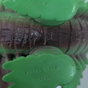 May include: A green and brown plastic monster figure with textured skin. The underside of the feet features molded text including BANDAI 1989 JAPAN on one foot and Japanese characters on the other, indicating the manufacturer and production year.