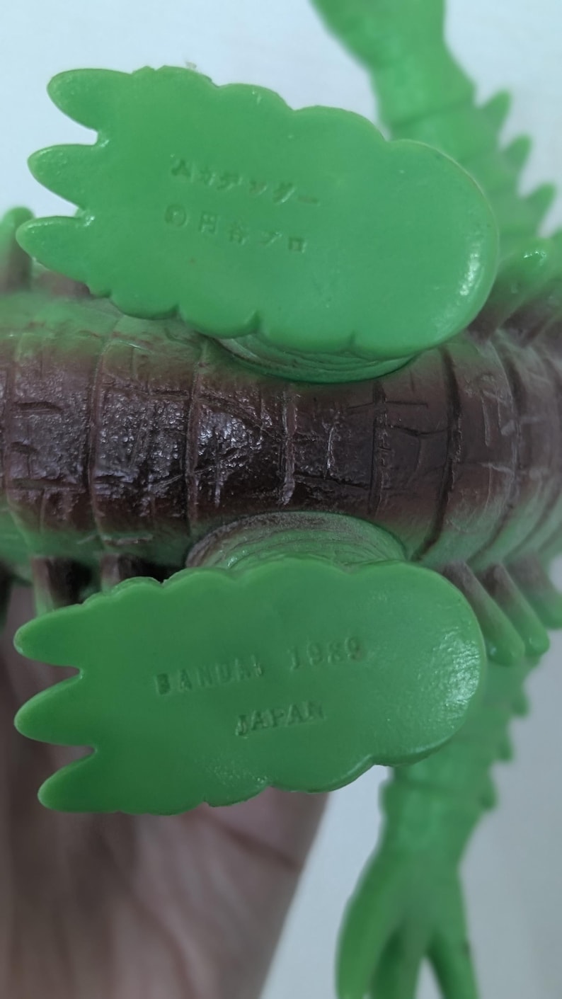 May include: A green and brown plastic monster figure with textured skin. The underside of the feet features molded text including BANDAI 1989 JAPAN on one foot and Japanese characters on the other, indicating the manufacturer and production year.