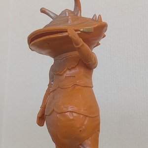 May include: An orange-colored figurine of a creature with a large, flat head and a segmented body. The figure is standing upright, with arms raised, and appears to be made of plastic or a similar material. The figurine has a unique, stylized design.