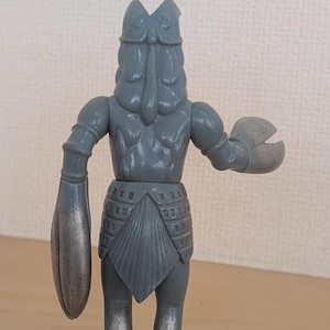 May include: A gray, vintage-style action figure of a creature with a crab-like claw and a large, armored arm. The figure has a detailed design, with a textured body and a helmet-like head. The figure is standing on a wooden surface.