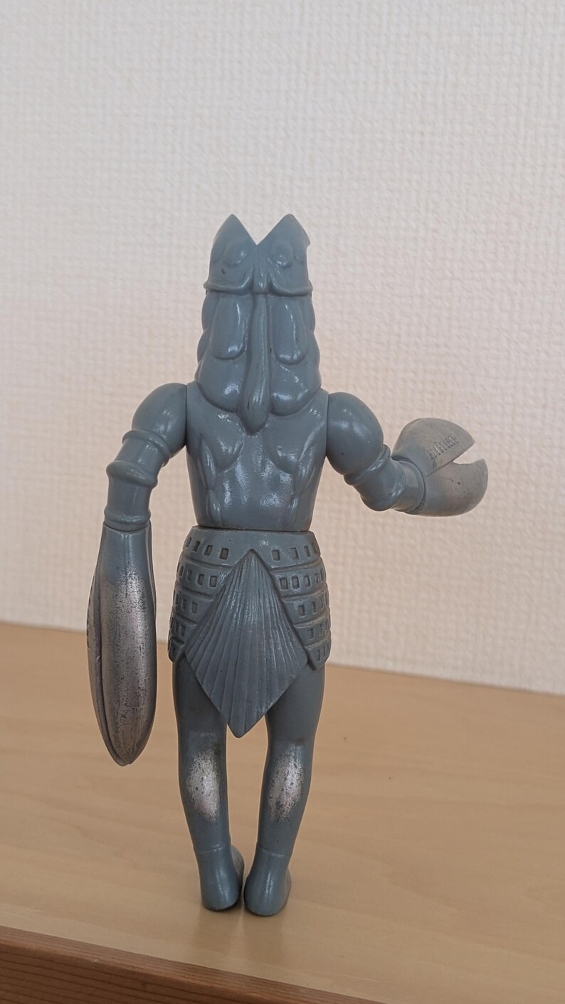 May include: A gray, vintage-style action figure of a creature with a crab-like claw and a large, armored arm. The figure has a detailed design, with a textured body and a helmet-like head. The figure is standing on a wooden surface.
