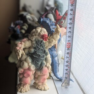 Op de afbeelding: Een beige en roze getextureerd monsterfiguur met een donkergrijze borstplaat en rode gezichtsdetails, staand naast een verticaal meetlint dat een hoogte van ongeveer 21,5 cm voor het wezen aangeeft.
