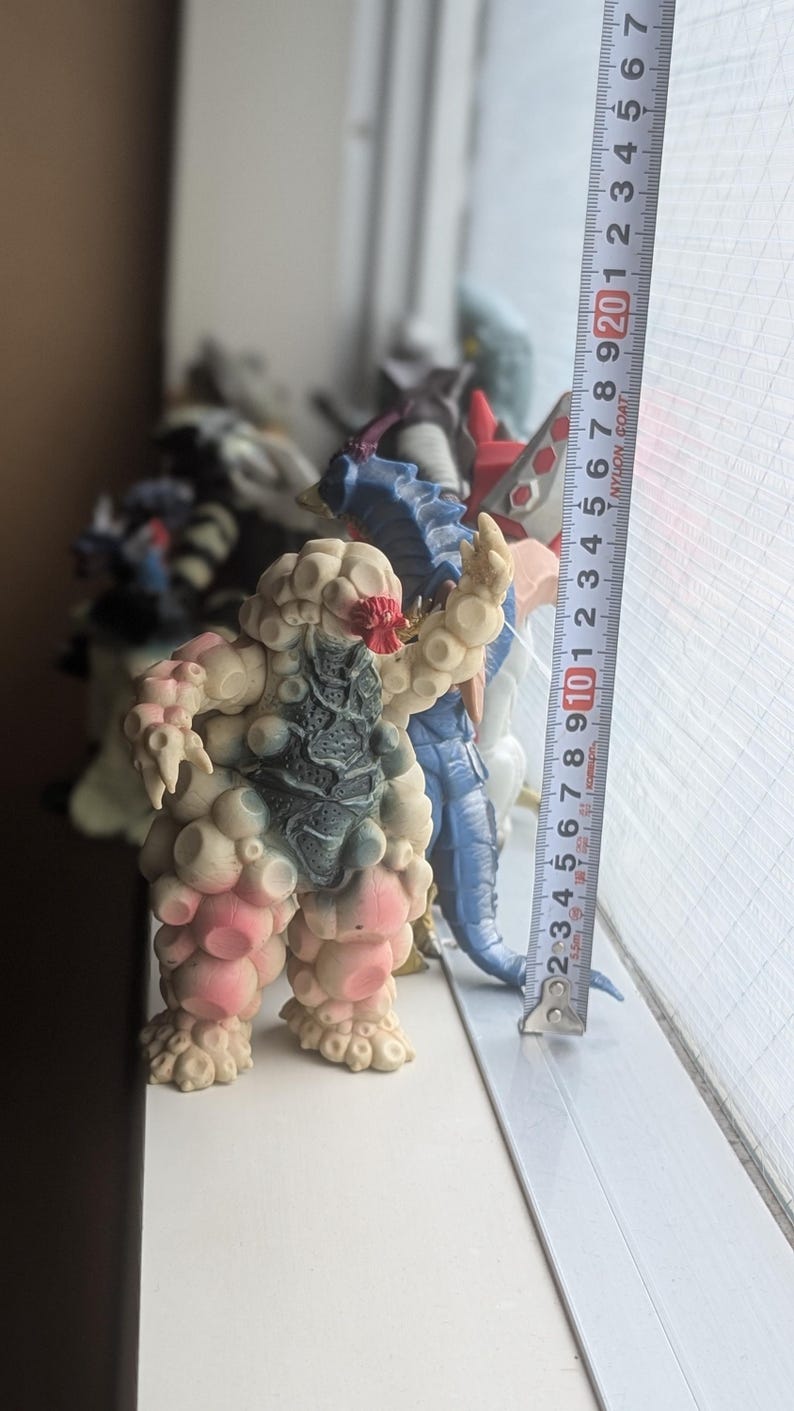 Op de afbeelding: Een beige en roze getextureerd monsterfiguur met een donkergrijze borstplaat en rode gezichtsdetails, staand naast een verticaal meetlint dat een hoogte van ongeveer 21,5 cm voor het wezen aangeeft.