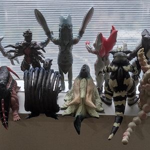 Op de afbeelding: Een verzameling van tien diverse monsteractiefiguren die in twee rijen staan. De figuren hebben verschillende vormen, texturen en kleuren, waaronder grijs, zwart, beige en blauw, met opvallende ledematen, staarten en stekels aan de achterkant.