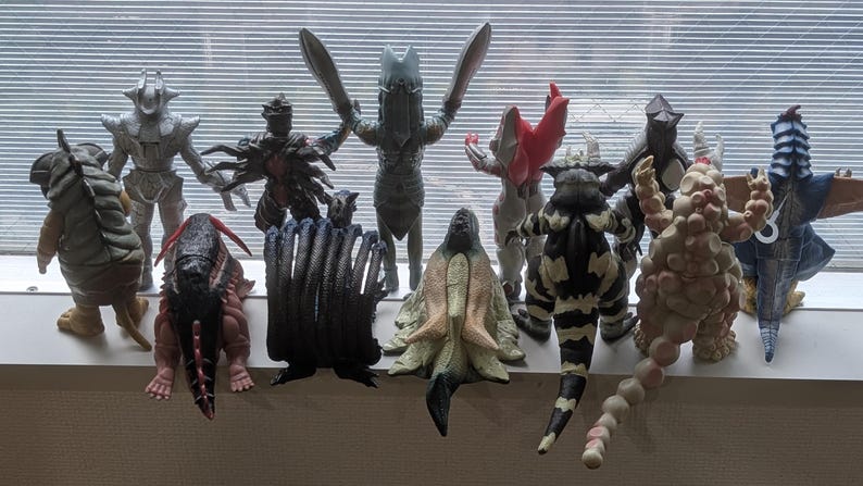 Op de afbeelding: Een verzameling van tien diverse monsteractiefiguren die in twee rijen staan. De figuren hebben verschillende vormen, texturen en kleuren, waaronder grijs, zwart, beige en blauw, met opvallende ledematen, staarten en stekels aan de achterkant.