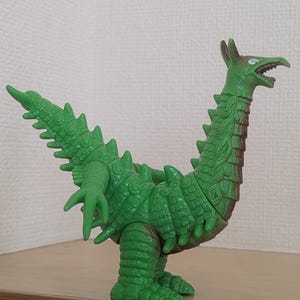May include: A green plastic monster figurine featuring a long neck, a pointed head with an open mouth, and a textured body covered in spikes. The creature has a long, spiked tail, two arms with three fingers each, and two thick legs.