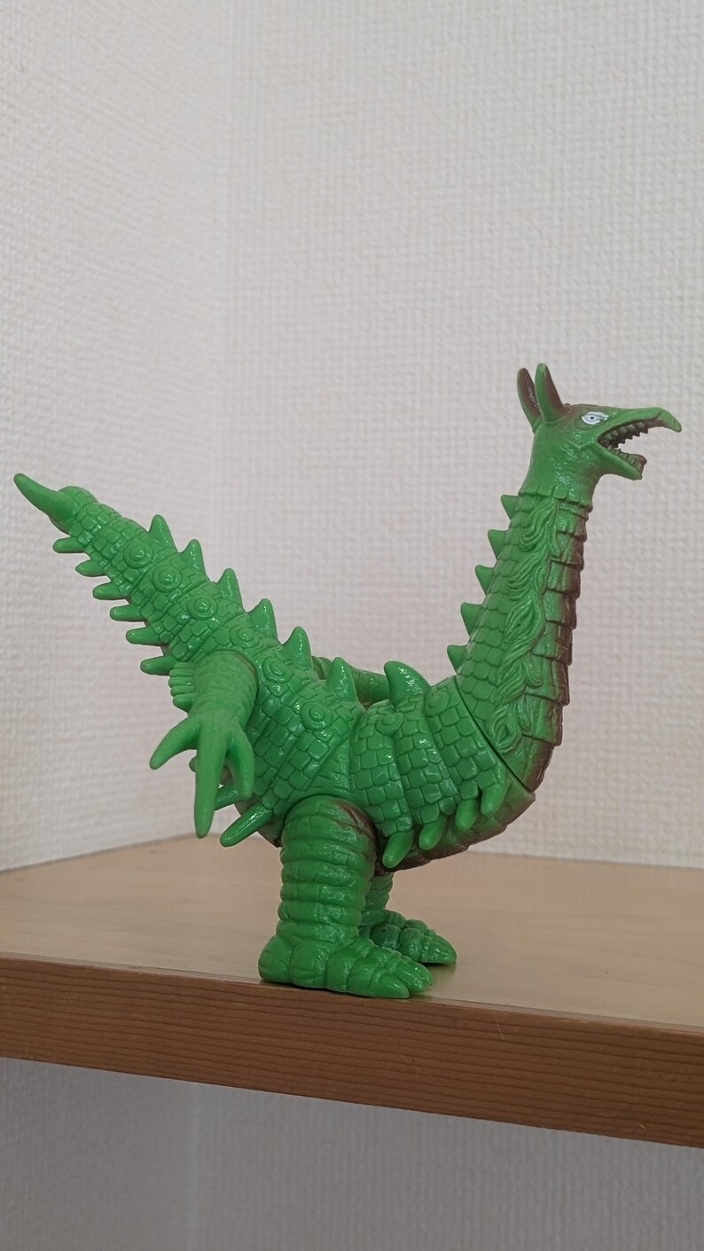 May include: A green plastic monster figurine featuring a long neck, a pointed head with an open mouth, and a textured body covered in spikes. The creature has a long, spiked tail, two arms with three fingers each, and two thick legs.