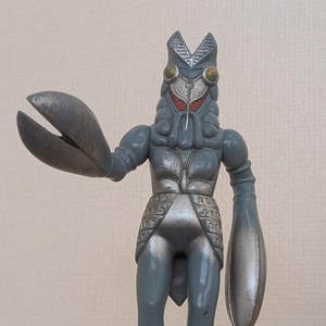 May include: A gray, articulated monster figure with a crab-like claw and a rounded arm. The figure has a detailed head with large yellow eyes and a red face. The figure is standing on a wooden surface.