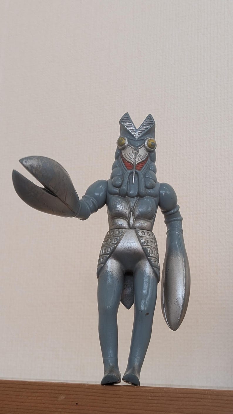 May include: A gray, articulated monster figure with a crab-like claw and a rounded arm. The figure has a detailed head with large yellow eyes and a red face. The figure is standing on a wooden surface.