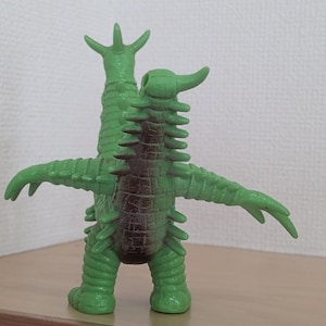 May include: A green monster figurine with two heads, multiple spikes along its back and arms, and a dark brown patch on its torso. The creature stands upright with its arms extended outward, featuring a textured, reptilian-like skin surface.